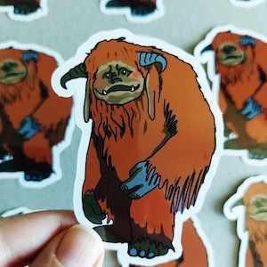 May include: A sticker featuring a cartoon illustration of a furry orange creature with blue horns and feet. The creature has a grumpy expression and is standing on all fours.