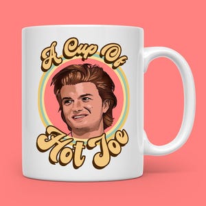 May include: White ceramic mug with a portrait of a man with brown hair, set within a retro design. The mug features the text "A Cup of Hot Joe" in a stylized font. The mug has a white handle and is set against a pink background.