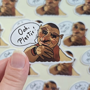 May include: A sticker featuring a cartoon character with a tan complexion, wearing a red vest and a bracelet. The character has a speech bubble that says, "Ooh, Plastic!" The sticker is held by a hand, with other similar stickers in the background.