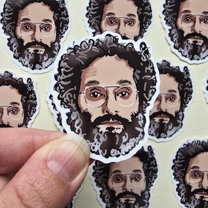 May include: A collection of stickers featuring a portrait of a man with glasses, a beard, and curly hair. The stickers are white with a detailed illustration in shades of brown, black, and gray. The stickers are held by a person's hand.