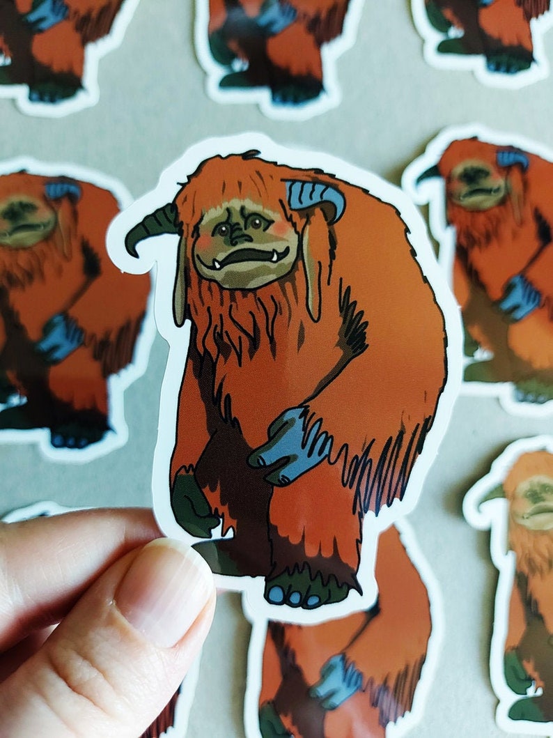 May include: A sticker featuring a cartoon illustration of a furry orange creature with blue horns and feet. The creature has a grumpy expression and is standing on all fours.