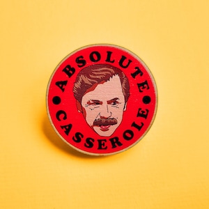 May include: A red and white round pin with a cartoon illustration of a man with a moustache and the text "ABSOLUTE CASSEROLE" in black.