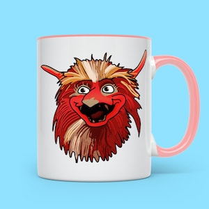 May include: White ceramic mug with a pink handle. The mug features a cartoon illustration of a red, furry monster with a wide, toothy grin.