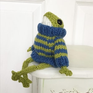 May include: A green knitted frog wearing a blue and green striped sweater. The frog is sitting on a white surface with its legs outstretched.