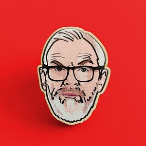 May include: A wooden pin featuring a cartoon illustration of a man with a white beard, grey hair, and glasses. The man is wearing a white shirt and has a serious expression.