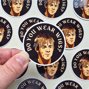 May include: Round stickers featuring a portrait of a person with light brown hair. The stickers are black with white text that reads "DO YOU WEAR WIGS?" The person's face is in shades of brown and yellow.