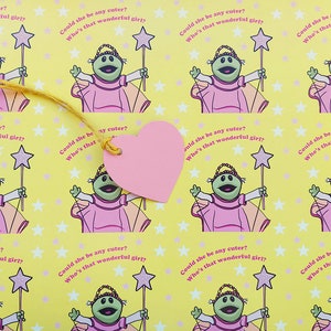 May include: A yellow background with a repeating pattern of a green puppet wearing a pink dress and holding a wand with a star on top. The text "Could she be any cuter? Who's that wonderful girl?" is repeated throughout the pattern. A pink heart-shaped tag is attached to the pattern with a yellow string.