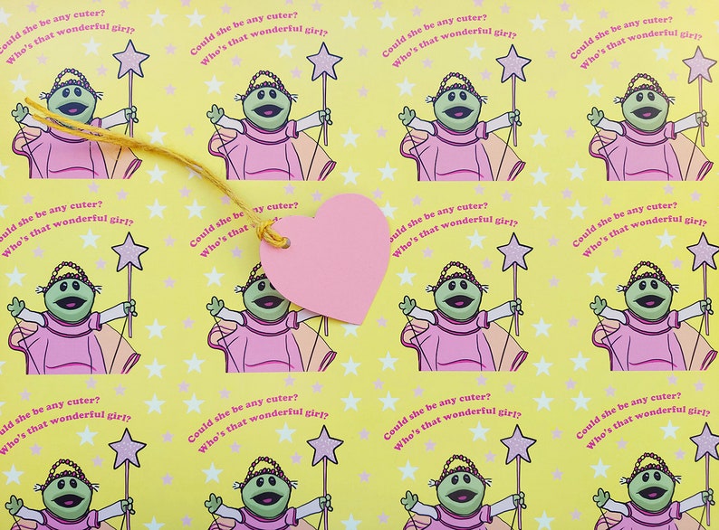 May include: A yellow background with a repeating pattern of a green puppet wearing a pink dress and holding a wand with a star on top. The text "Could she be any cuter? Who's that wonderful girl?" is repeated throughout the pattern. A pink heart-shaped tag is attached to the pattern with a yellow string.
