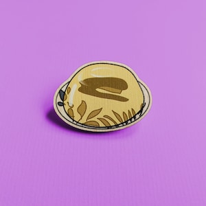 May include: A wooden pin shaped like a plate with a yellow pudding with a brown topping on it.