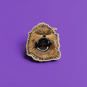 Peut inclure: Un écusson représentant une créature de dessin animé rugissante avec une fourrure marron et beige, une bouche ouverte et des dents pointues. L'écusson a une bordure claire et est posé sur un fond violet.