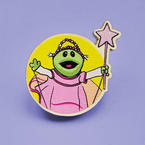 May include: A green frog wearing a pink dress and a crown holds a pink star-shaped wand. The frog is on a yellow background.