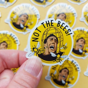 May include: A set of yellow and black stickers featuring the text "Not the Bees!" and a cartoon image of a man with bees around his head. The man is wearing a black shirt and has a scared expression on his face.