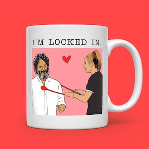 May include: White ceramic mug with the text "I'M LOCKED IN." The mug features a cartoon illustration of a man and woman with a heart and a red spoon. The background is pink.
