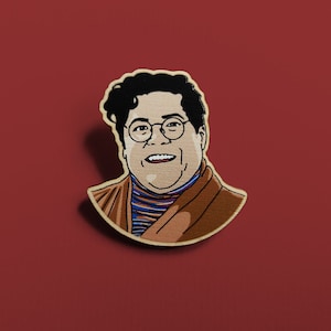May include: A wooden pin featuring a cartoon illustration of a person with brown hair, wearing glasses, a brown sweater, and a blue and white striped shirt. The person is smiling.