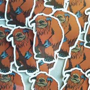 May include: A pile of orange and blue fuzzy creature stickers. The stickers are of a creature with large, furry ears, a wide mouth, and a long, furry tail. The stickers are all facing the same direction.