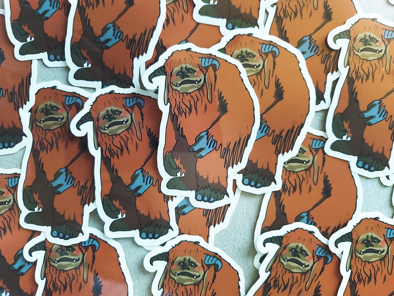 May include: A pile of orange and blue fuzzy creature stickers. The stickers are of a creature with large, furry ears, a wide mouth, and a long, furry tail. The stickers are all facing the same direction.