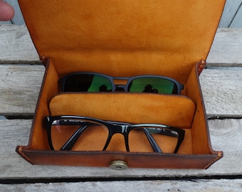 May include: A brown leather glasses case with two pairs of glasses inside. The top pair is sunglasses with green lenses, and the bottom pair is clear glasses with black frames.