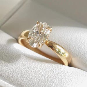 May include: A gold ring with a large, oval-cut diamond set in a classic, polished band. The ring is displayed in a white, textured jewelry box, creating a contrast between the bright diamond and the gold band.