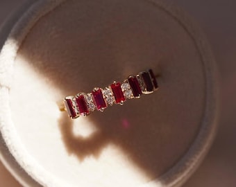 May include: A gold ring featuring alternating rectangular red gemstones and small white gemstones. The ring is set against a soft, neutral background, highlighting the vibrant colors of the stones. The ring is a piece of jewelry.