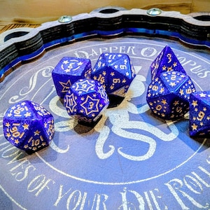 May include: A set of blue and gold polyhedral dice with star patterns, displayed on a blue surface with white text. The dice are on a wooden tray with a decorative edge, ideal for tabletop gaming.
