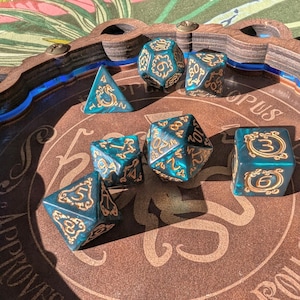 May include: A wooden dice tray with a blue and white border. The tray has the words "S. Approuve Rolls Corpus" printed on it. There are six teal and gold dice with black numbers on them inside the tray.
