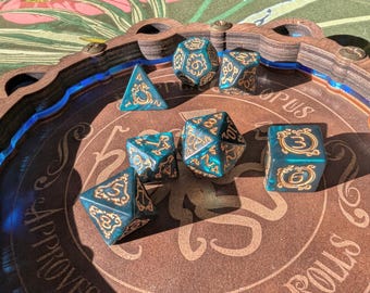 May include: A wooden dice tray with a blue and white border. The tray has the words "S. Approuve Rolls Corpus" printed on it. There are six teal and gold dice with black numbers on them inside the tray.