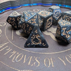 May include: A set of black and gold dice with intricate designs. The dice are on a brown leather surface with gold lettering that reads "Approves of Your Choice".