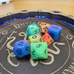 May include: A selection of multi-sided dice in a range of colours, including blue, green, and orange, are displayed on a brown tray. The dice are of various types, such as D6, D10, and D20, and have black numbering. The tray has decorative elements.