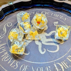 May include: A selection of clear dice containing yellow duck figures, displayed on a purple surface. The dice are in different shapes, such as a d20, d10, and d6. The surface features the text "Super Octopus Approves of Your Die Rolls."