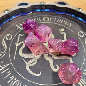 May include: A selection of translucent pink and purple dice with white numbers sits on a dark grey dice tray. The tray has a decorative border and the words "Super Octopus Approved Rolls" are printed on it.