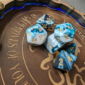 May include: A wooden dice tray with a brown interior and blue LED trim, holding several polyhedral dice. The dice are white and teal with gold numbering. The tray has gold text that reads "Approves of Your October".