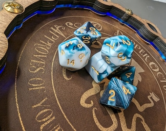 May include: A wooden dice tray with a brown interior and blue LED trim, holding several polyhedral dice. The dice are white and teal with gold numbering. The tray has gold text that reads "Approves of Your October".
