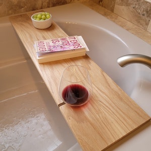 May include: A wooden bathtub tray holds a glass of red wine, a bowl of green grapes, and a book titled "It Ends With Us". The tray is made of light-colored wood and spans the width of the bathtub.