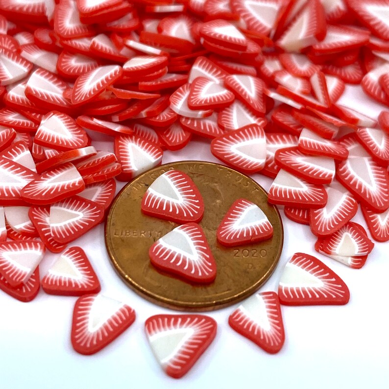 May include: A large pile of red and white strawberry slice shaped polymer clay charms. The charms are scattered on a white surface with a US penny for scale.