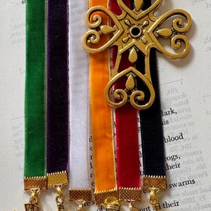 May include: A collection of velvet ribbon bookmarks in green, purple, white, orange, red, and black. Each bookmark has a gold-colored cross charm. A larger, ornate gold cross is attached to the top. The bookmarks are on a book page.