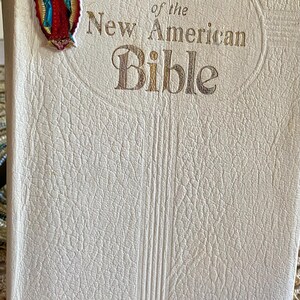 May include: A white leather-bound Bible with gold lettering. The title reads "Saint Joseph Edition of the New American Bible". A blue ribbon bookmark with a silver charm and a colorful image of the Virgin Mary is attached to the book.