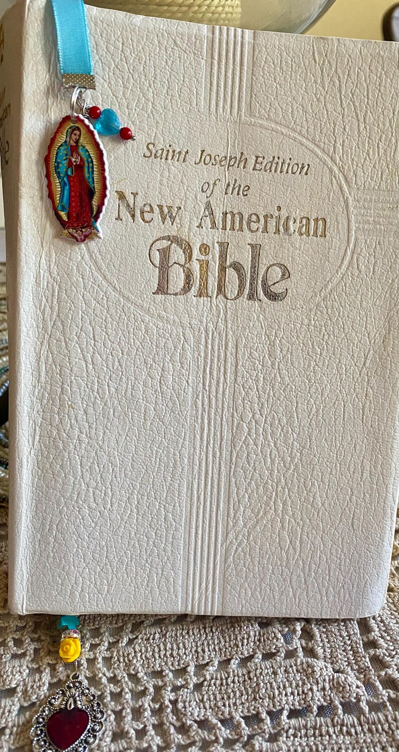 May include: A white leather-bound Bible with gold lettering. The title reads "Saint Joseph Edition of the New American Bible". A blue ribbon bookmark with a silver charm and a colorful image of the Virgin Mary is attached to the book.