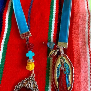 May include: A blue velvet ribbon necklace with a silver heart charm, a turquoise cross charm, and a charm with a picture of the Virgin Mary. The charms are attached to the ribbon with silver rings. The necklace is on a red, white, and green striped background.