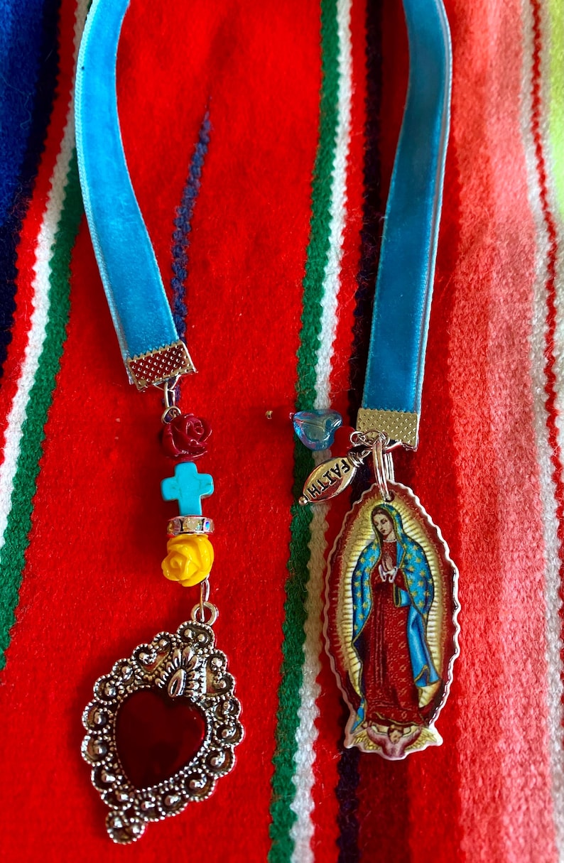 May include: A blue velvet ribbon necklace with a silver heart charm, a turquoise cross charm, and a charm with a picture of the Virgin Mary. The charms are attached to the ribbon with silver rings. The necklace is on a red, white, and green striped background.