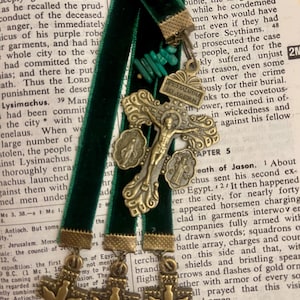 Pu&ograve; includere: Quattro segnalibri in velluto verde con dettagli in metallo dorato e bronzo. Ogni segnalibro presenta una croce con una colomba e un piccolo ciondolo in metallo. I segnalibri sono appoggiati su una pagina di un libro con del testo.