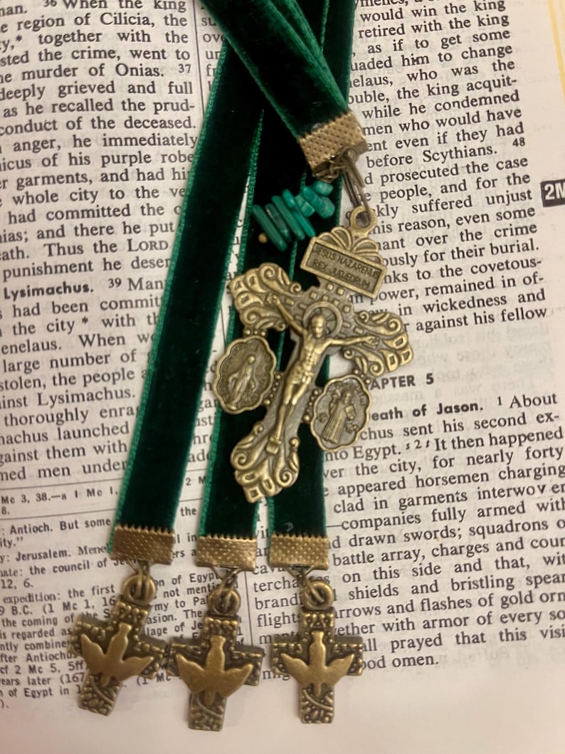 Pu&ograve; includere: Quattro segnalibri in velluto verde con dettagli in metallo dorato e bronzo. Ogni segnalibro presenta una croce con una colomba e un piccolo ciondolo in metallo. I segnalibri sono appoggiati su una pagina di un libro con del testo.