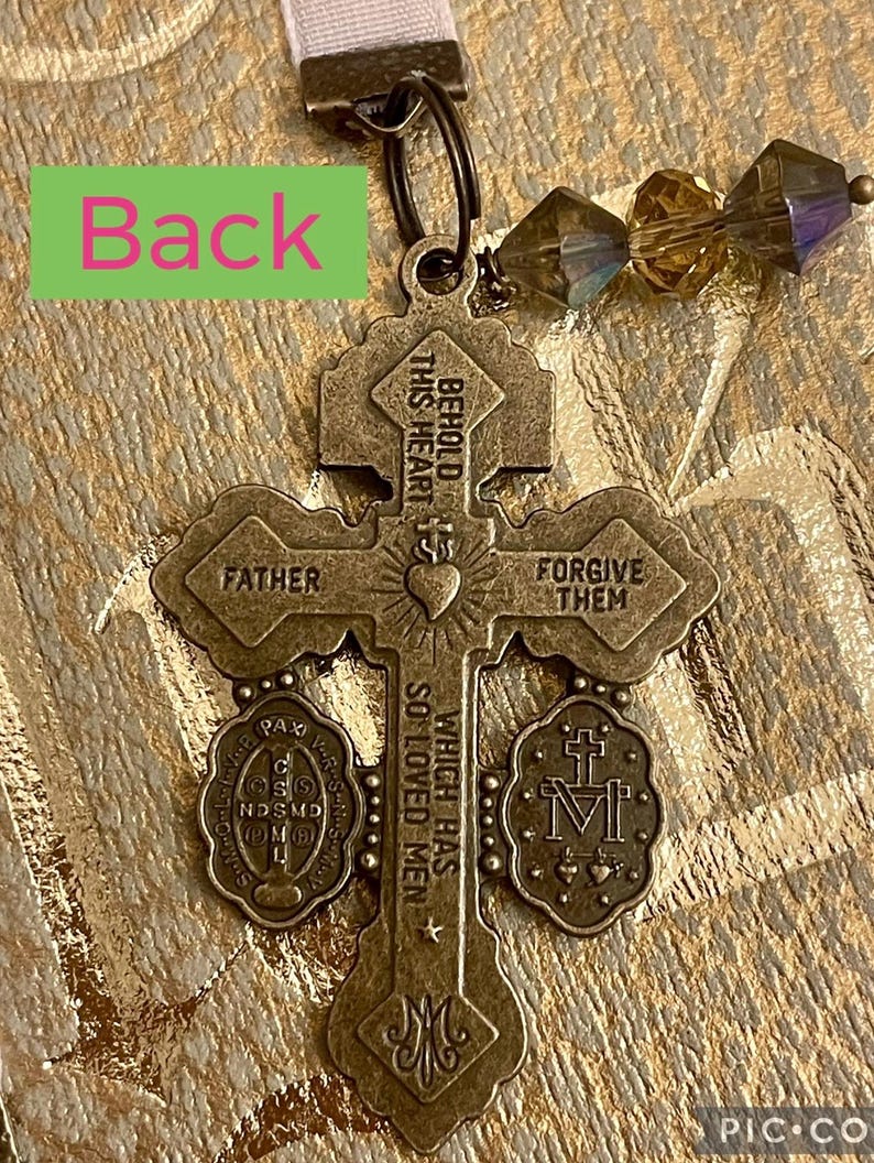 Pu&ograve; includere: Un ciondolo a forma di croce di colore bronzo con le parole "Behold this heart which has so loved men" e "Forgive them" sul davanti. Il retro del ciondolo ha le parole "Father" e un piccolo ciondolo rotondo con le parole "Pax Vobis" e una croce.