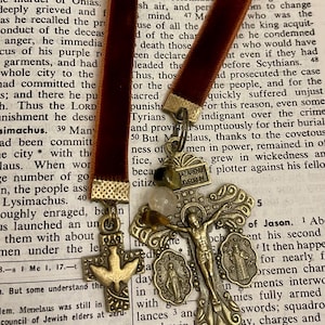 Pu&ograve; includere: Primo piano di un nastro di velluto marrone con una variet&agrave; di ciondoli religiosi attaccati. I ciondoli includono una croce con una figura di Ges&ugrave; Cristo, una colomba e un piccolo ciondolo rotondo con le parole "LAZARUS JUDAEORUM".