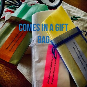 May include: Three different colored fabric gift bags with tags that say "Bible Bookmark Handmade for you to keep your favorite scriptures marked". The bags are arranged on a wooden surface. The text "COMES IN A GIFT BAG" is superimposed on the image.
