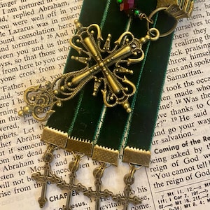 May include: Four green velvet ribbon bookmarks with gold metal cross charms and a single red and green glass bead charm. The bookmarks are on a page of a book with the text "Four Sayings of Jesus" and "Scandals will inevitably come."