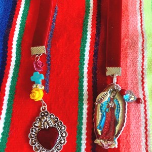 May include: Two red velvet ribbon bookmarks with silver charms. One charm features a red heart with silver filigree and the other charm features a depiction of the Virgin Mary with the text "Virgen" in silver lettering.