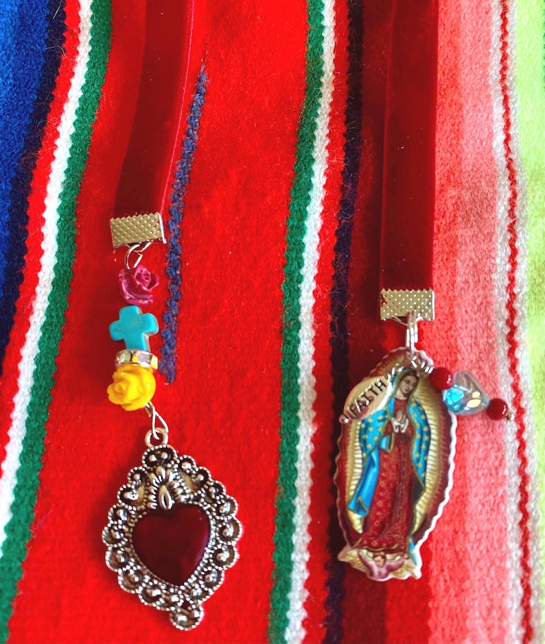 May include: Two red velvet ribbon bookmarks with silver charms. One charm features a red heart with silver filigree and the other charm features a depiction of the Virgin Mary with the text "Virgen" in silver lettering.