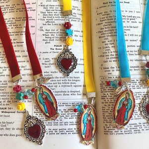 May include: Three colorful bookmarks with religious imagery. The bookmarks feature a red velvet ribbon, a turquoise blue ribbon, and a yellow ribbon. Each bookmark has a metal heart charm with a red glass center. The bookmarks also feature a small image of the Virgin Mary. The bookmarks are on top of open books with text.