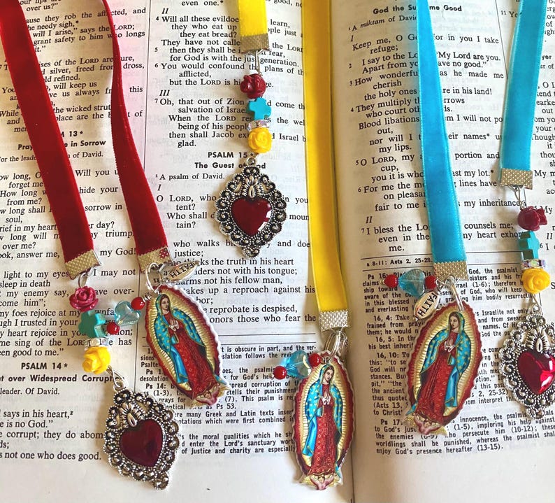 May include: Three colorful bookmarks with religious imagery. The bookmarks feature a red velvet ribbon, a turquoise blue ribbon, and a yellow ribbon. Each bookmark has a metal heart charm with a red glass center. The bookmarks also feature a small image of the Virgin Mary. The bookmarks are on top of open books with text.
