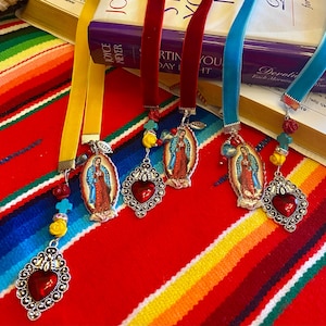 May include: Four colorful ribbon bookmarks with silver charms featuring an image of the Virgin Mary, a red heart, and various beads. The bookmarks are displayed on a colorful striped fabric.